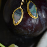 A pair of gold drop earrings with teardrop-shaped iridescent opal gemstones, displayed on a dark plum fruit with a leaf in the foreground.