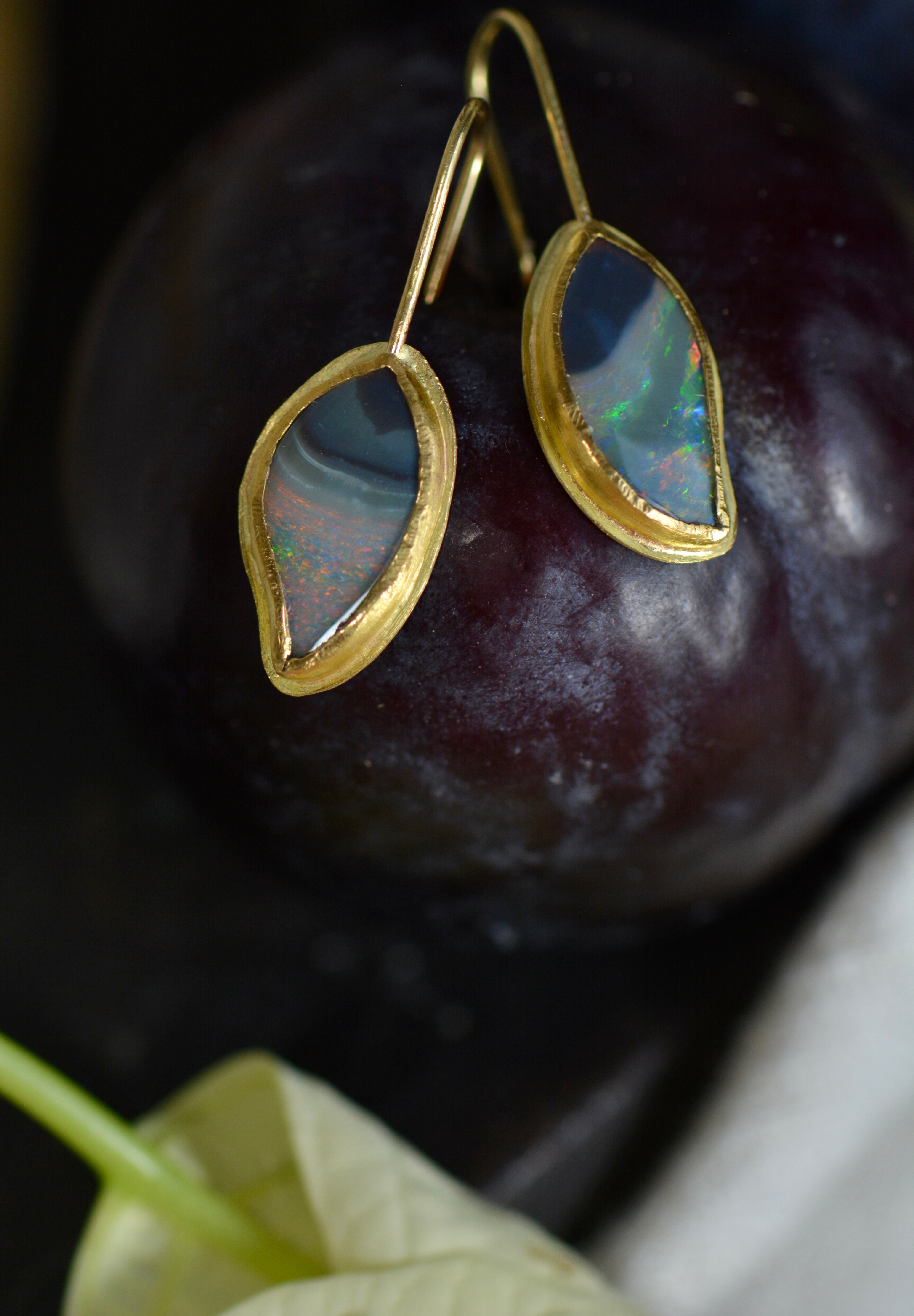 A pair of gold drop earrings with teardrop-shaped iridescent opal gemstones, displayed on a dark plum fruit with a leaf in the foreground.