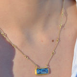 A close-up of a person's neck and chest wearing a delicate gold chain necklace with a rectangular blue stone pendant.