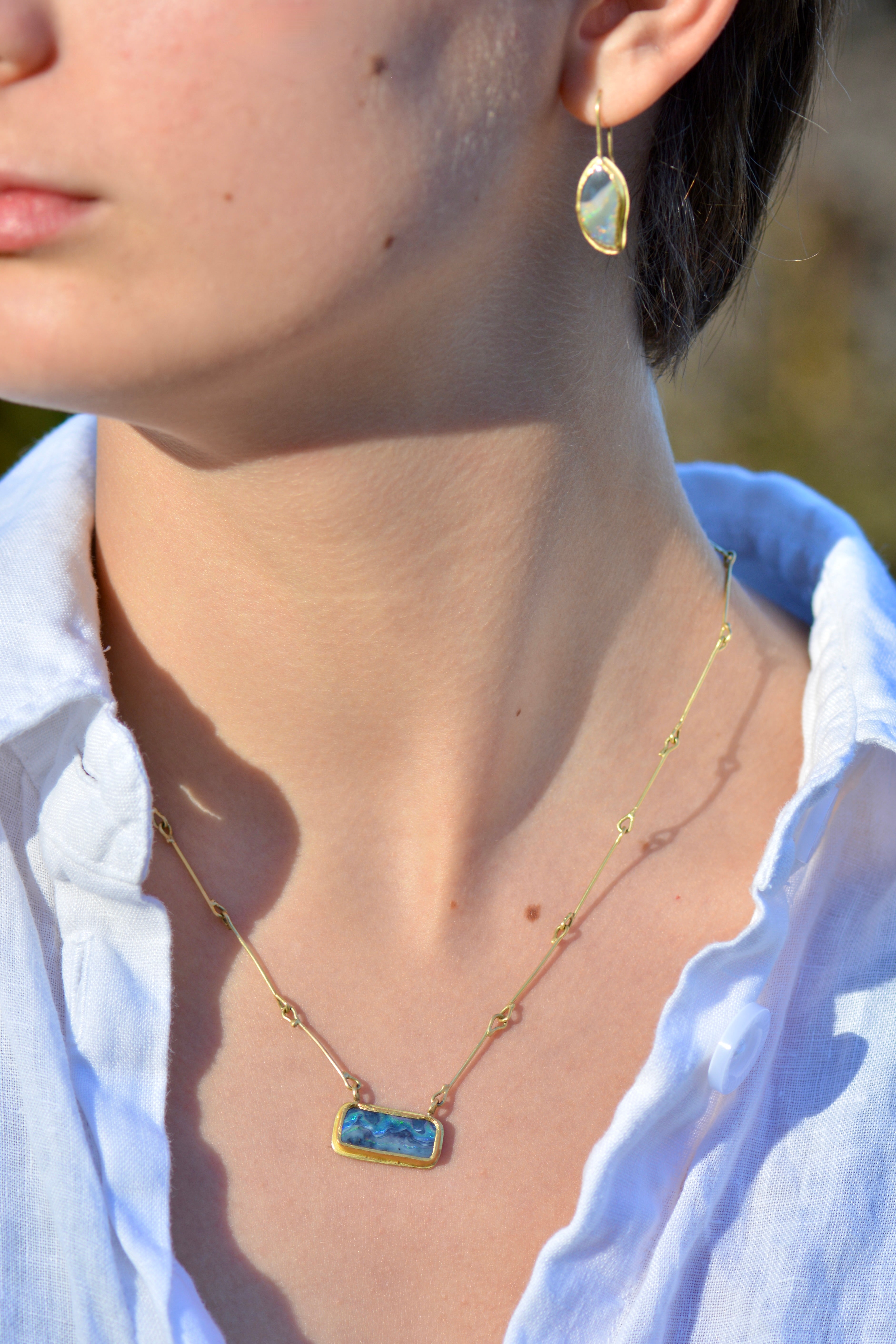 A close-up lifestyle shot of a woman wearing a gold and opal necklace with a blue rectangular stone pendant and a matching gold drop earring.
