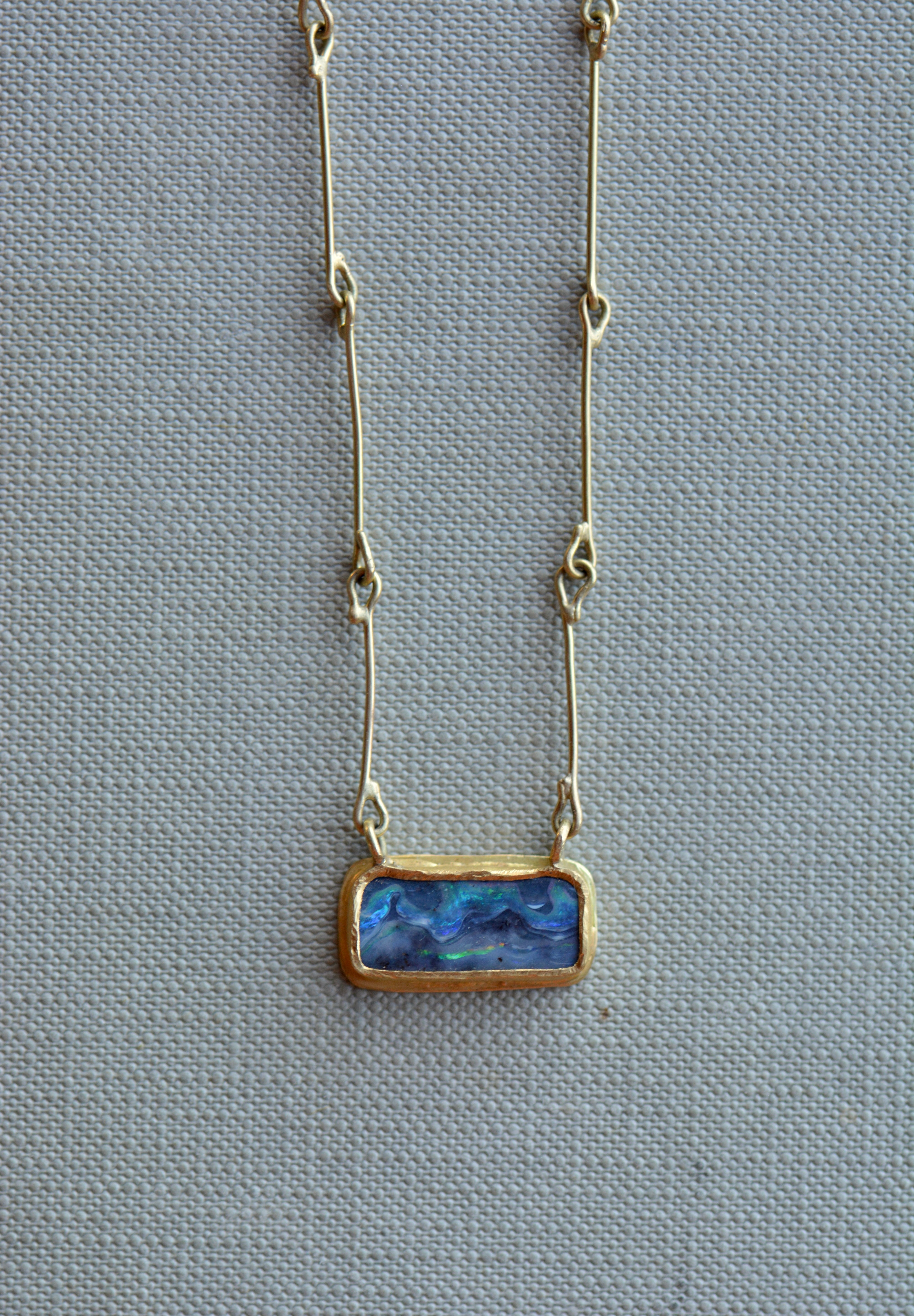 A delicate gold chain necklace featuring a rectangular opal pendant with a textured, shimmering blue stone, displayed on a neutral grey textured fabric background.