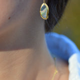 A close-up lifestyle shot of a person wearing a delicate gold dangle earring with an organic, irregularly shaped iridescent opal stone.