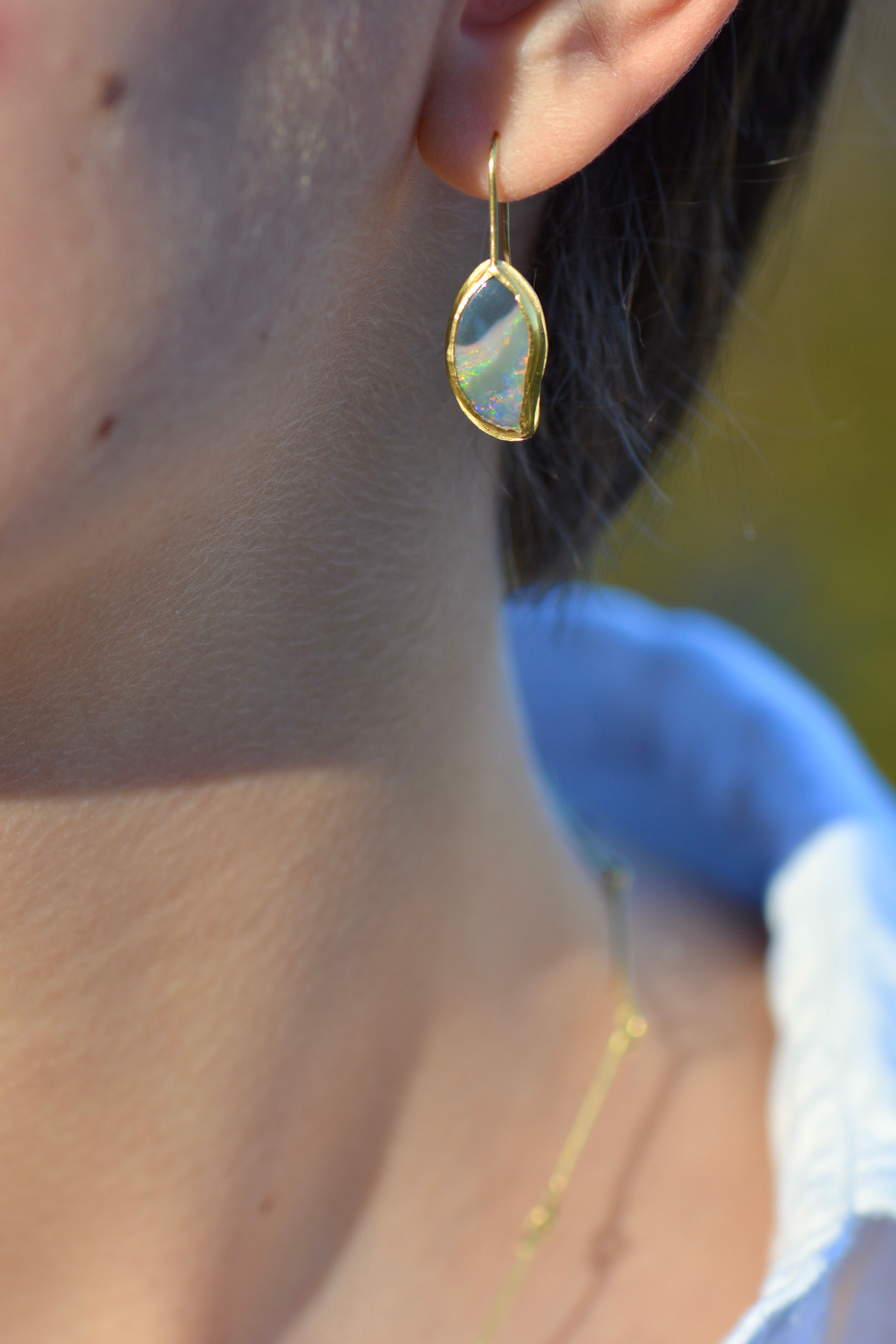 A close-up lifestyle shot of a person wearing a delicate gold dangle earring with an organic, irregularly shaped iridescent opal stone.