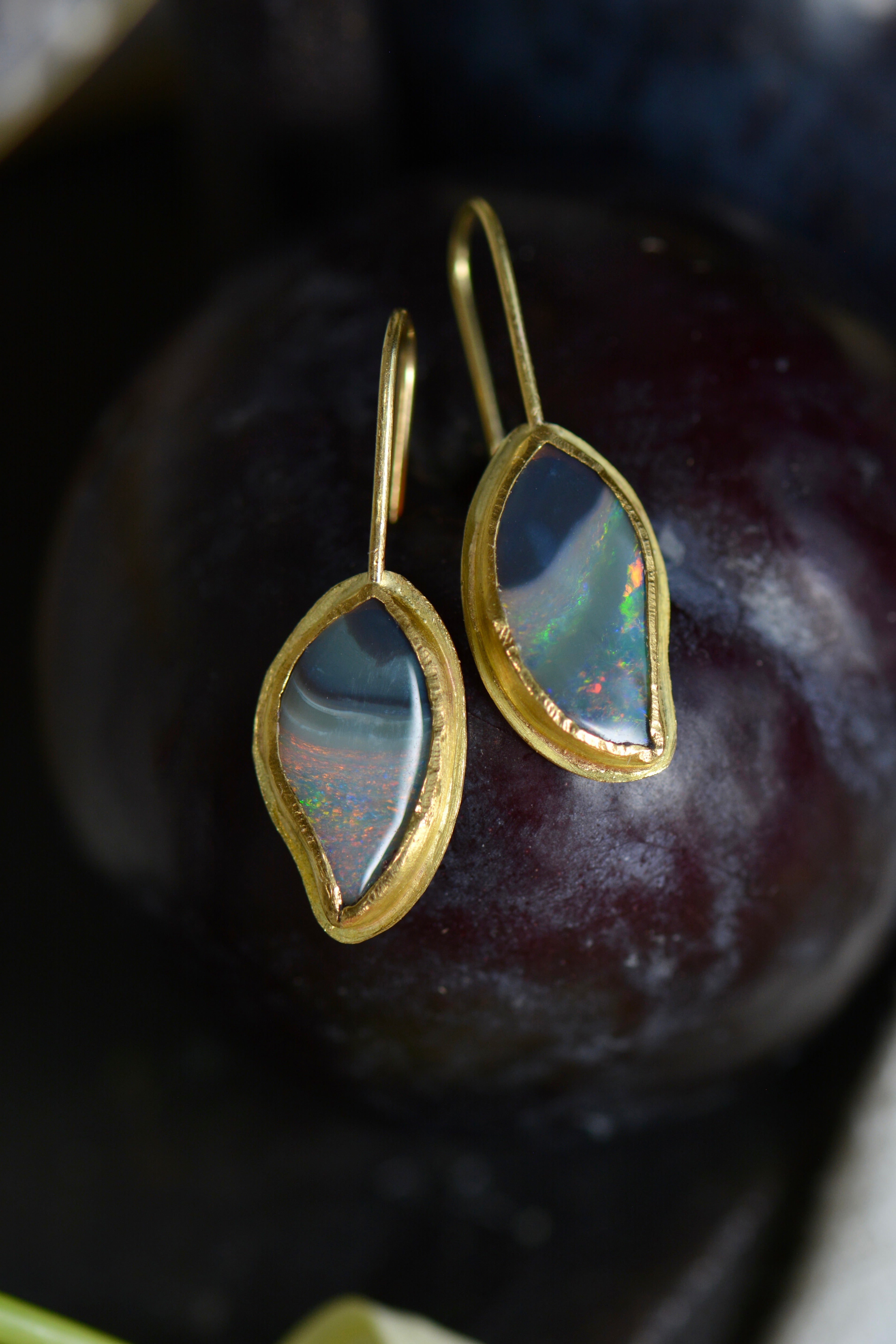 A pair of gold drop earrings with curvy marquise-shaped iridescent opal gemstones, resting on a dark, textured surface.