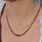 Close-up of a person wearing two delicate beaded necklaces: one in pale pink and cream pearl beads and one in warm pinkish-purple translucent beads, against a white linen blouse.