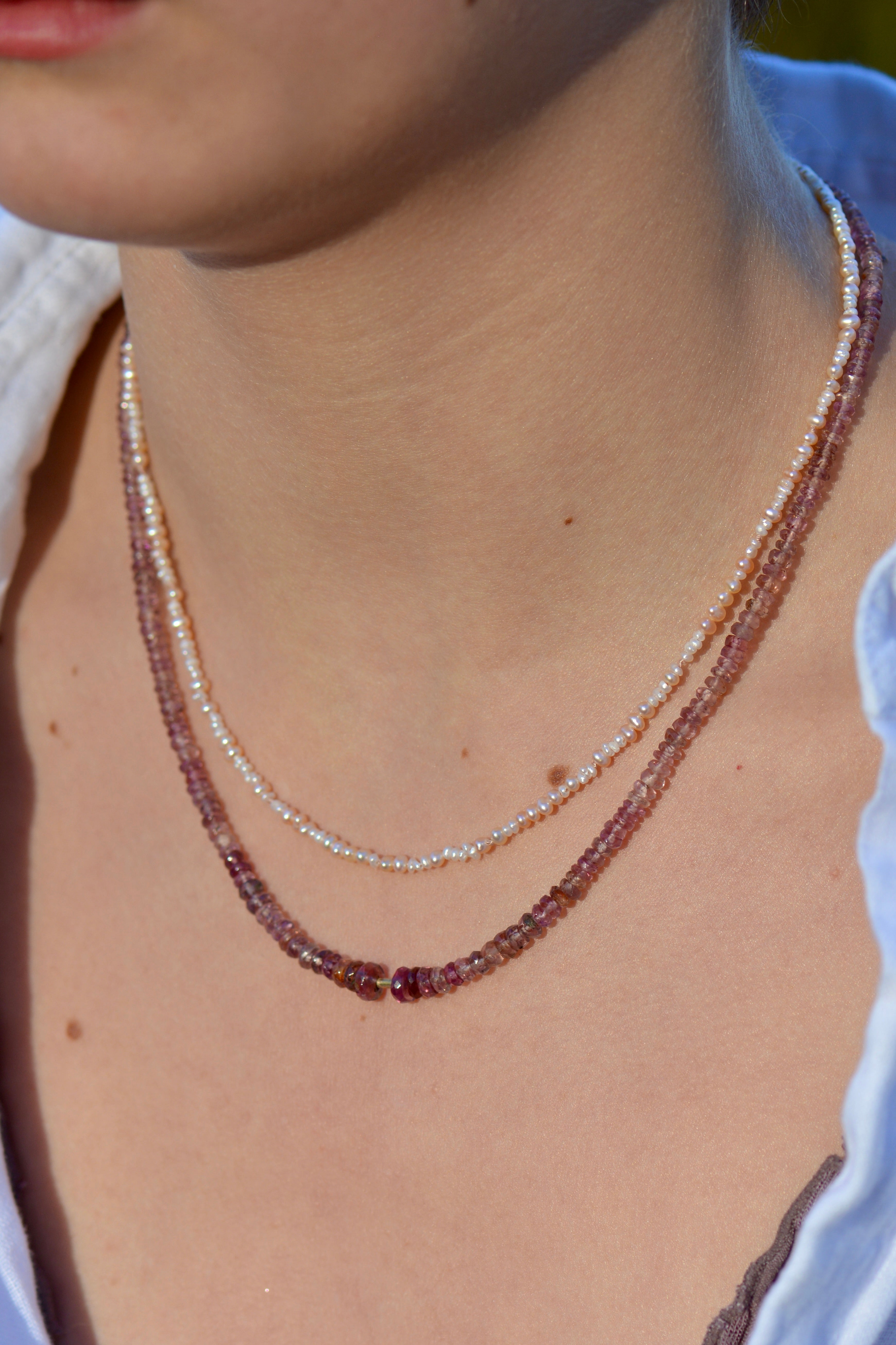 Close-up of a person wearing two delicate beaded necklaces: one in pale pink and cream pearl beads and one in warm pinkish-purple translucent beads, against a white linen blouse.