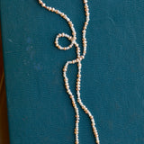 A strand of seed pearls in cream and pale pink with a gold clasp is shown on a teal backdrop.