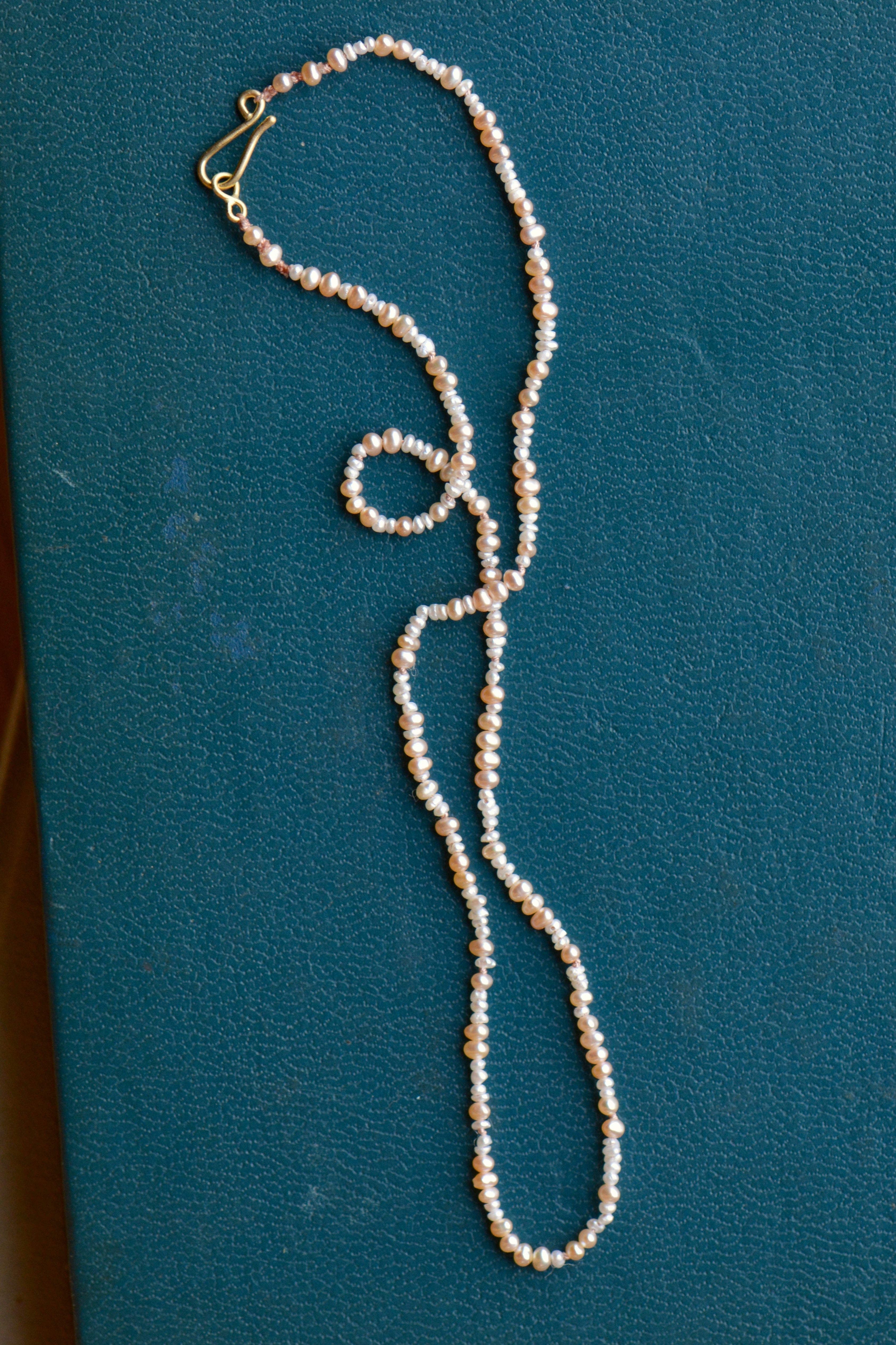 A strand of seed pearls in cream and pale pink with a gold clasp is shown on a teal backdrop.