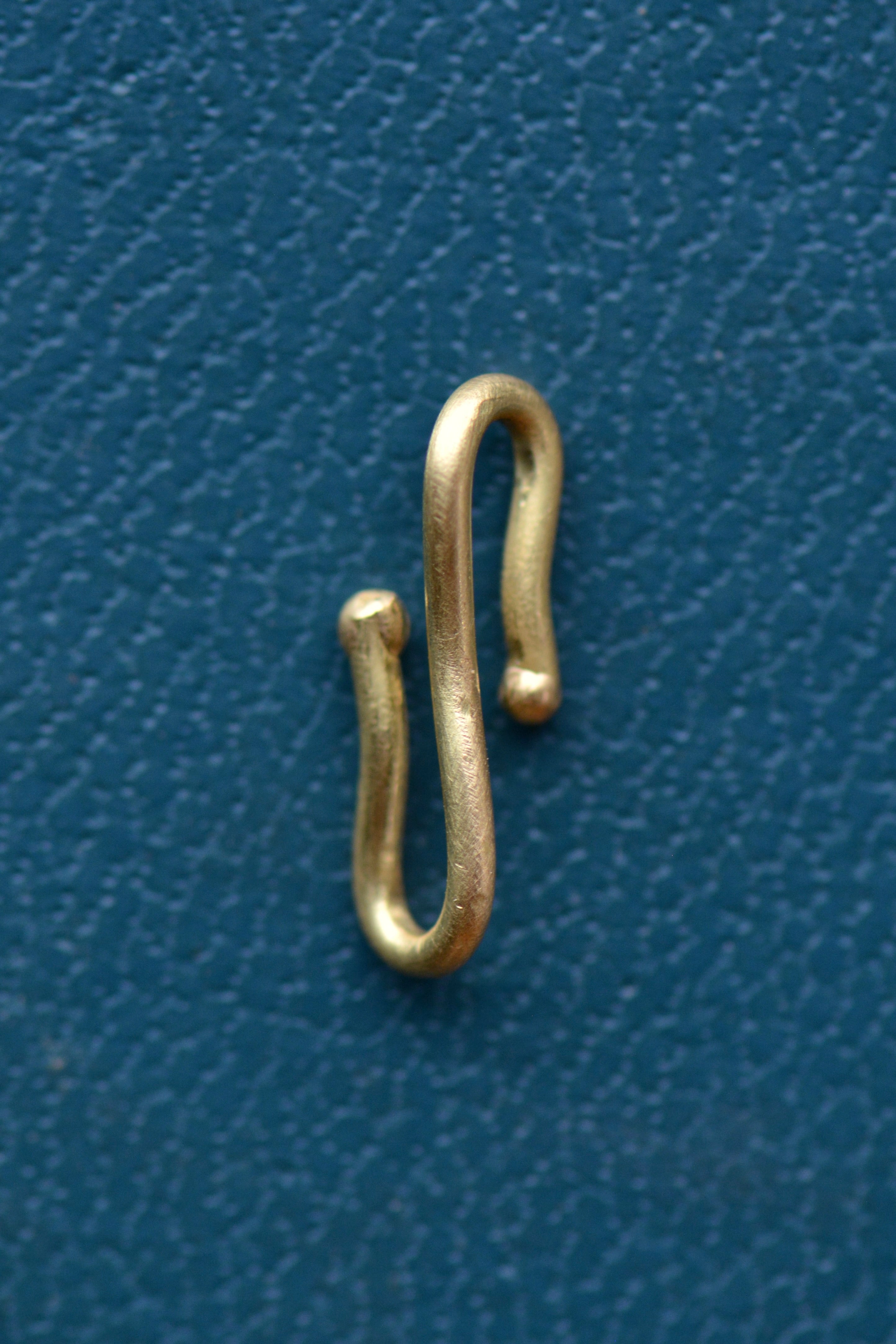 A gold charm connector hook sits on a teal blue textured backdrop.