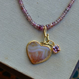 A gold heart pendant necklace with a striped orange dendritic agate stone and a small purple gemstone charm, displayed on a textured beige fabric surface.