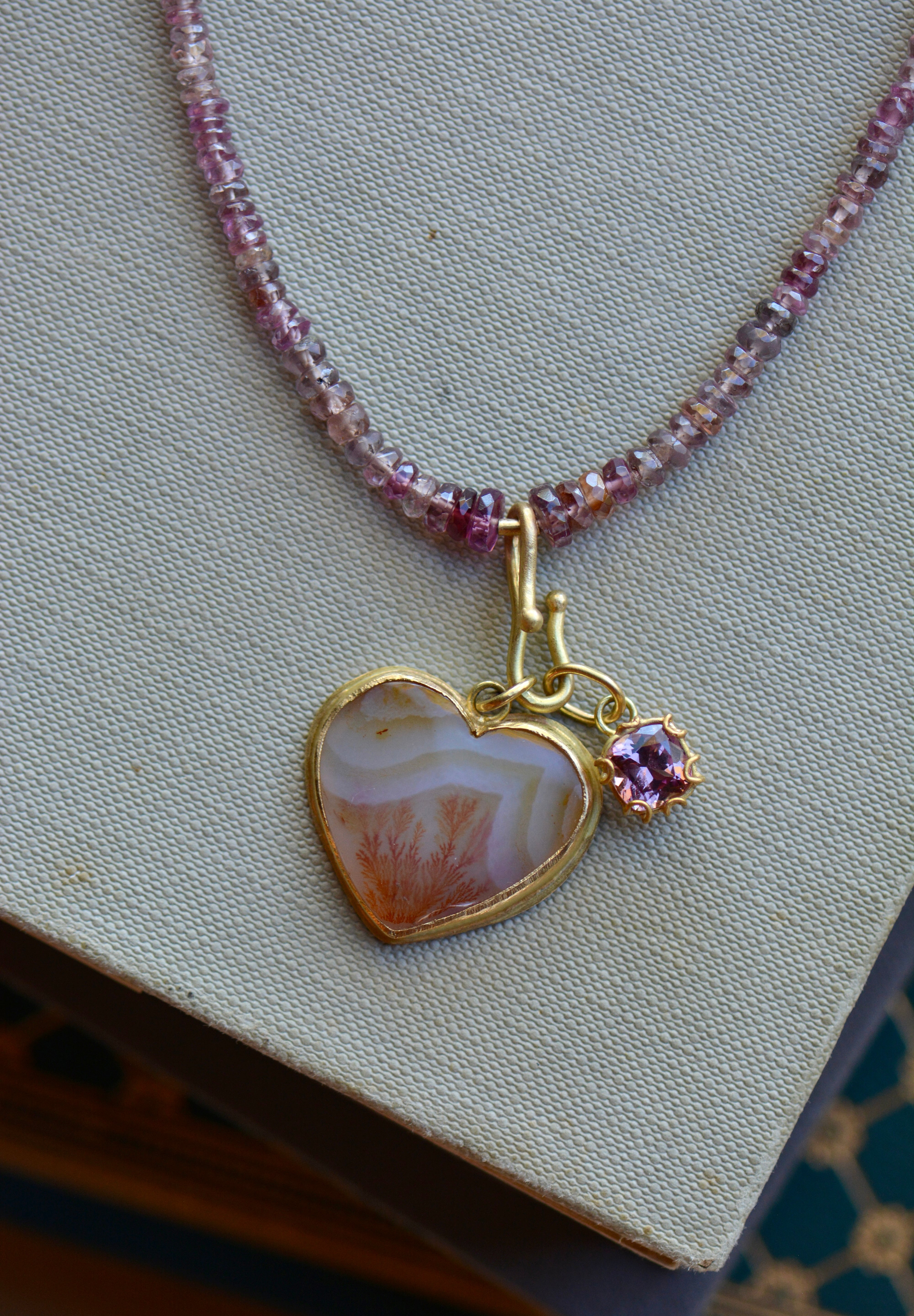 A gold heart pendant necklace with a striped orange dendritic agate stone and a small purple gemstone charm, displayed on a textured beige fabric surface.