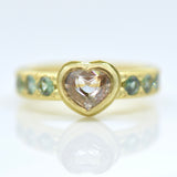 A gold ring featuring a heart-shaped translucent pale pink gemstone in the centre, with small blue gemstones set along the band, shown on a reflective white surface.