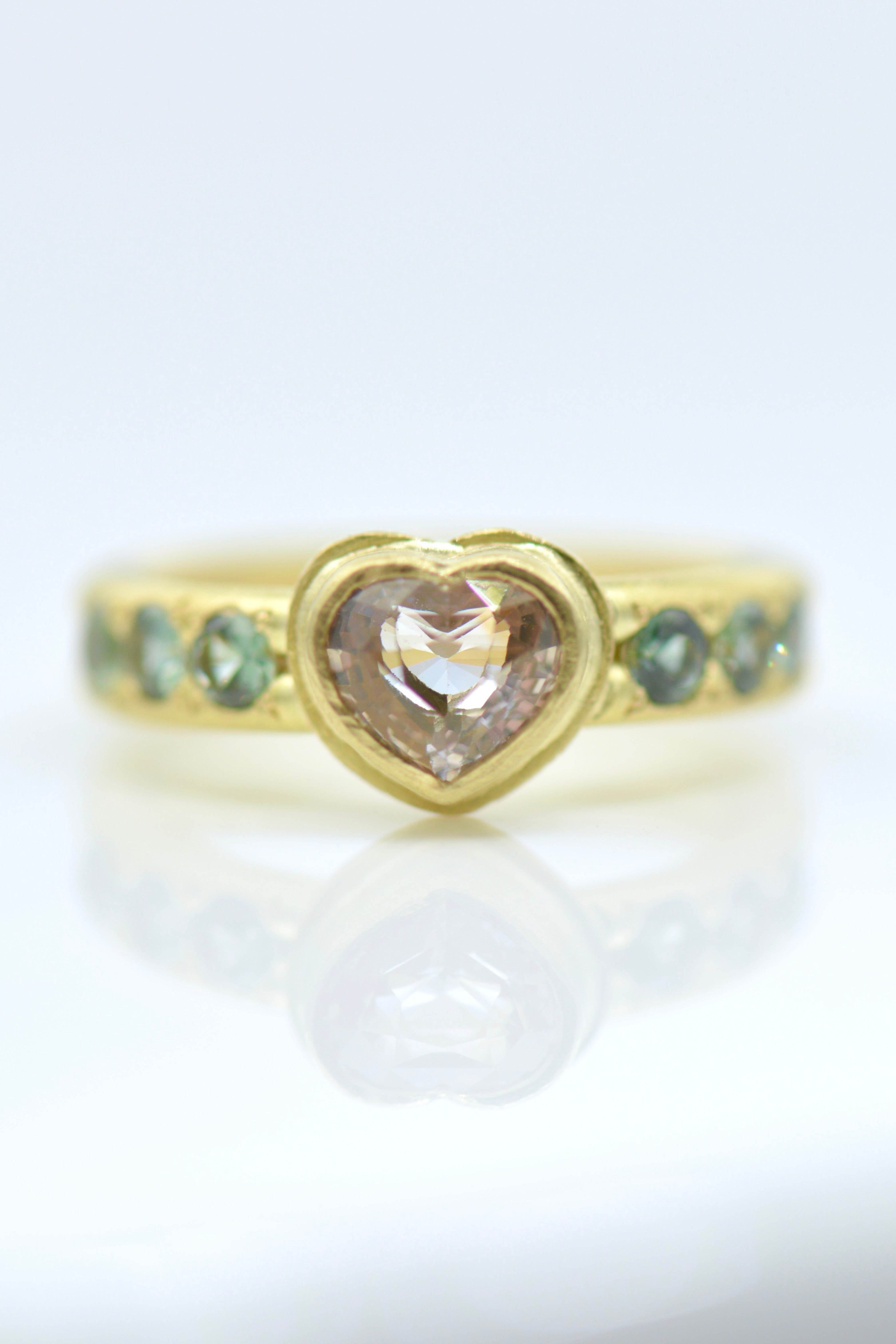 A gold ring featuring a heart-shaped translucent pale pink gemstone in the centre, with small blue gemstones set along the band, shown on a reflective white surface.