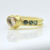 A yellow gold ring featuring a heart-shaped clear gemstone and three small round blue gemstones on each side of the band, displayed on a reflective white surface.