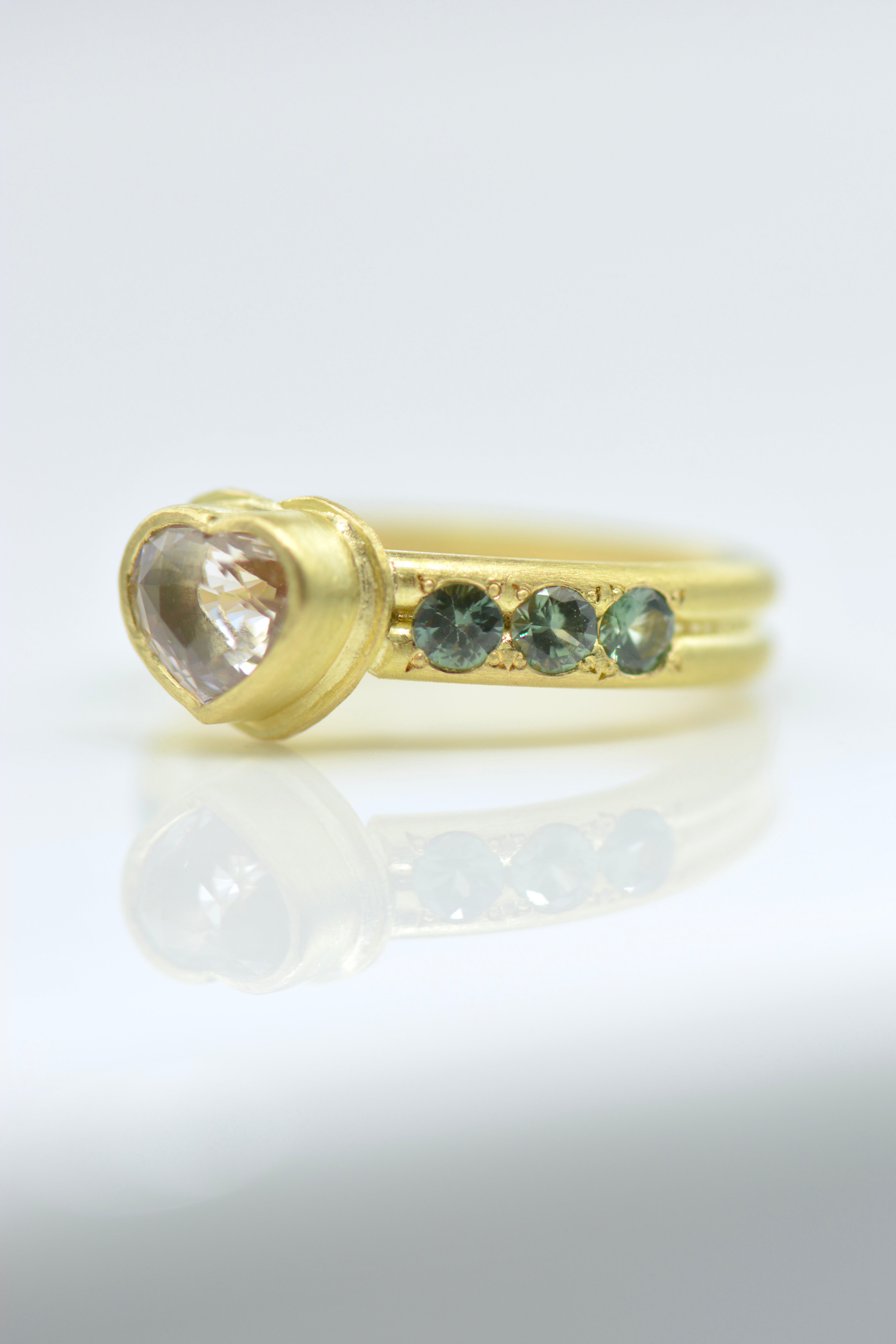 A yellow gold ring featuring a heart-shaped clear gemstone and three small round blue gemstones on each side of the band, displayed on a reflective white surface.