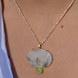 A close-up of a delicate pearl necklace worn on a person's neck, featuring a small beaded strand with a large oval white dendritic agate pendant featuring fine gray botanical patterns, and a small green opal stone set below in gold.