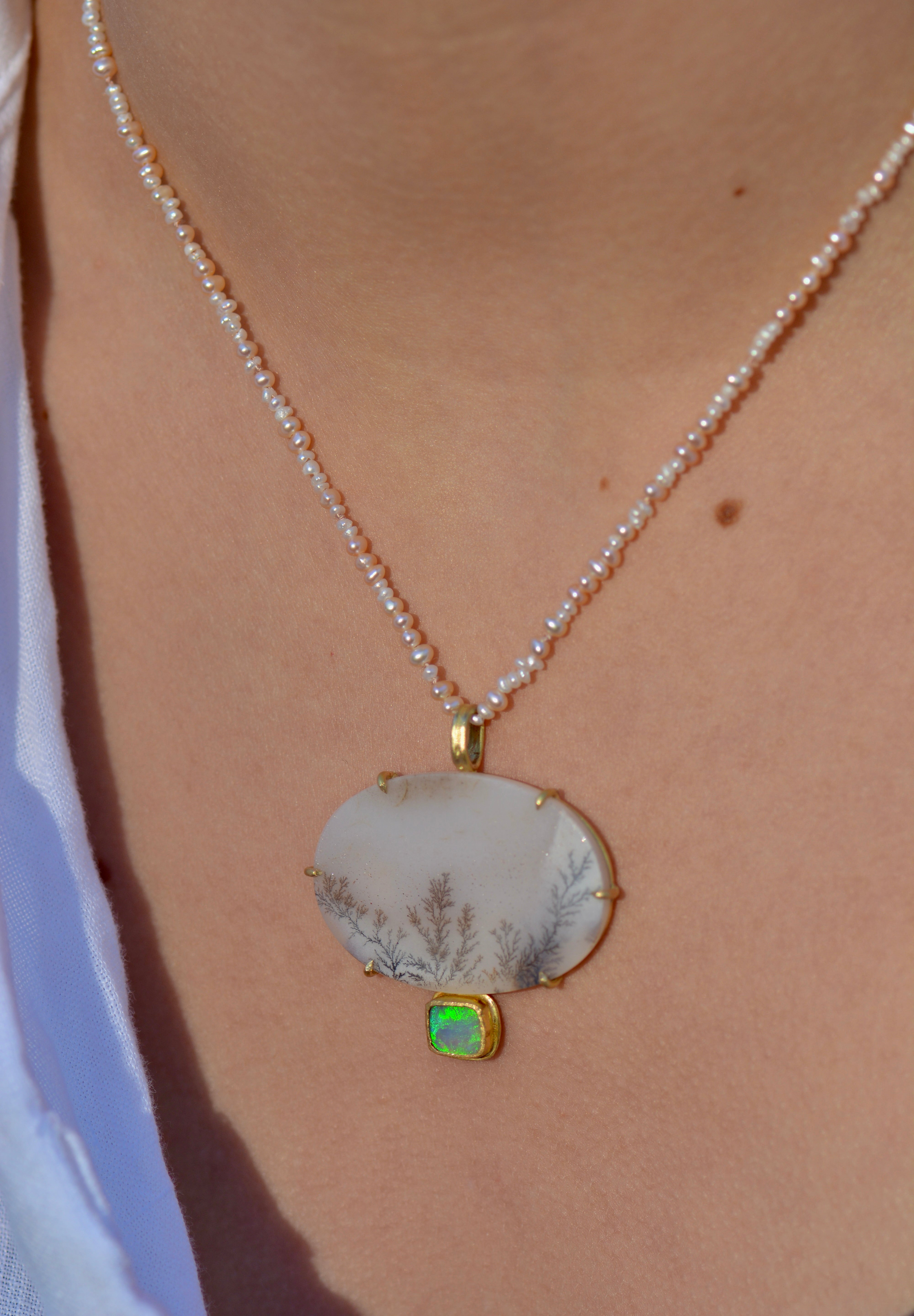 A close-up of a delicate pearl necklace worn on a person's neck, featuring a small beaded strand with a large oval white dendritic agate pendant featuring fine gray botanical patterns, and a small green opal stone set below in gold.