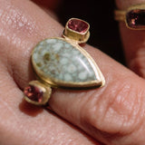 Close-up of a person's hands wearing multiple gold rings with colorful gemstones, including a prominent green turquoise and red stones, against a dark brown textured background.