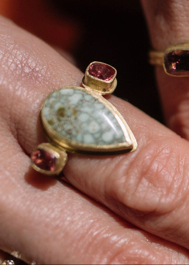 Close-up of a person's hands wearing multiple gold rings with colorful gemstones, including a prominent green turquoise and red stones, against a dark brown textured background.