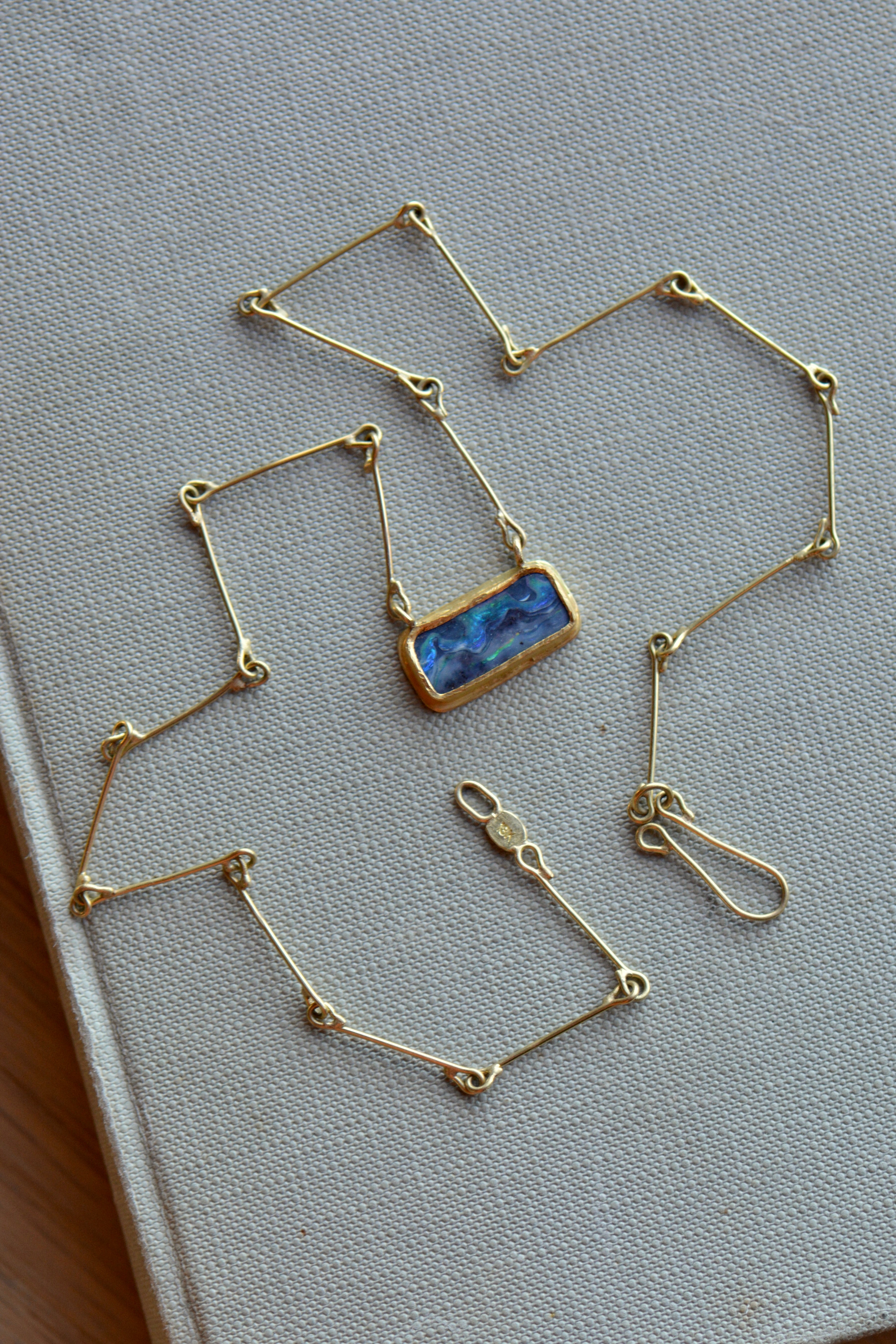 A gold geometric link chain necklace with a rectangular blue marbled opal pendant, displayed on a textured light grey book cover.