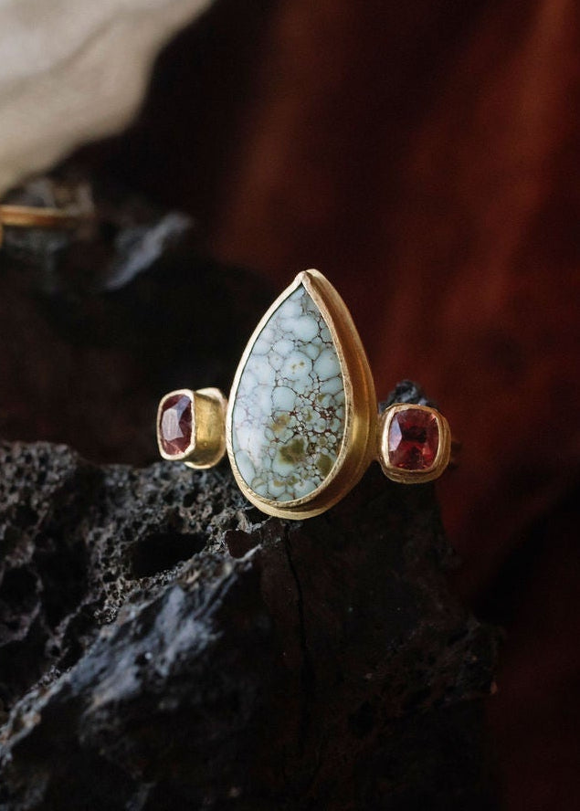 A styled jewelry arrangement featuring a gold ring with red and green stones, displayed on a textured black rock with a white fabric and dark red velvet background.