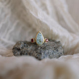 A gold ring with a teardrop-shaped light blue-green stone and small red gemstones on either side, displayed on a dark grey volcanic rock with a soft beige fabric background.