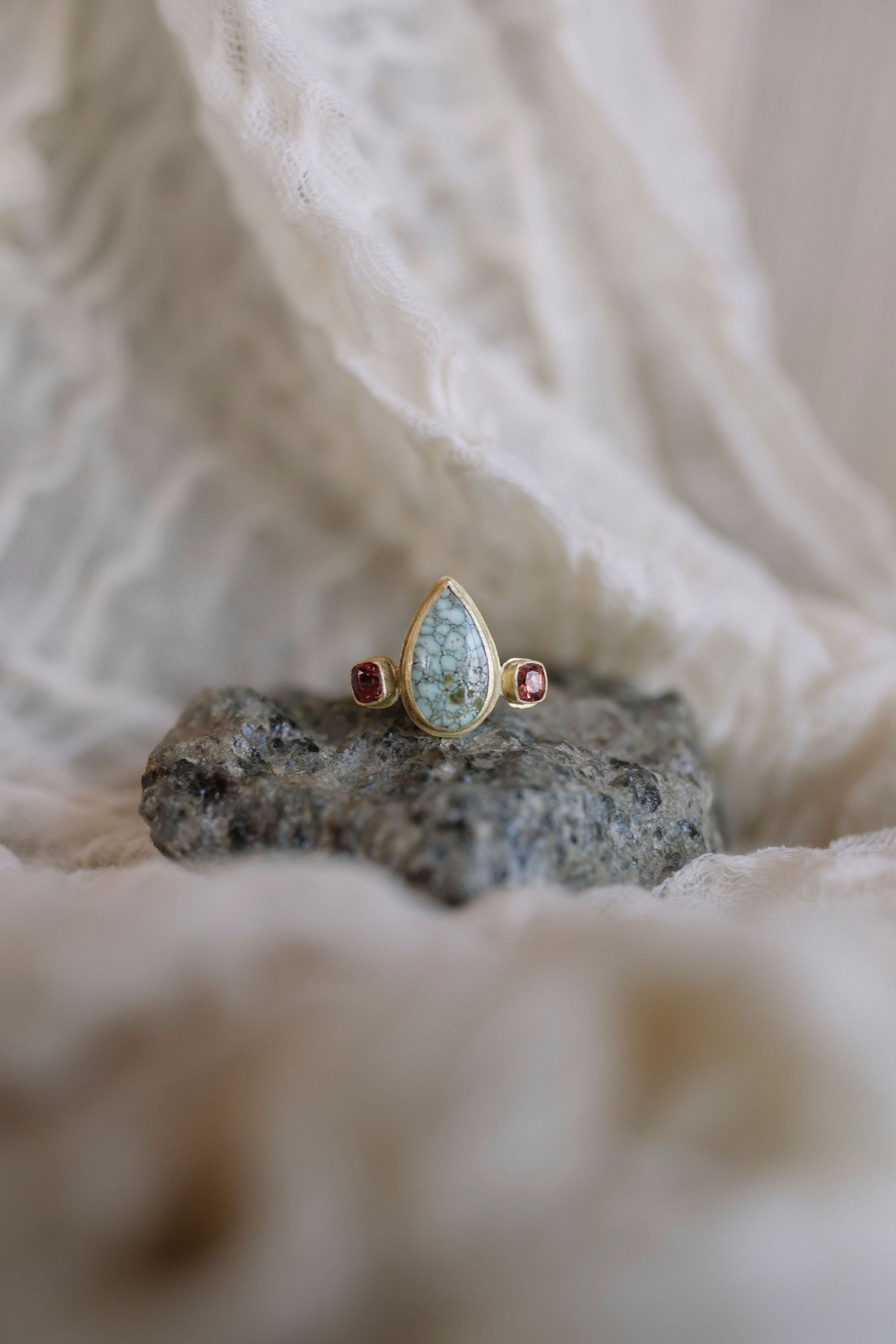 A gold ring with a teardrop-shaped light blue-green stone and small red gemstones on either side, displayed on a dark grey volcanic rock with a soft beige fabric background.