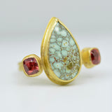 A gold ring featuring a large teardrop-shaped light green stone with a brown matrix pattern, flanked by two small faceted red gemstones, set against a neutral light gray background.