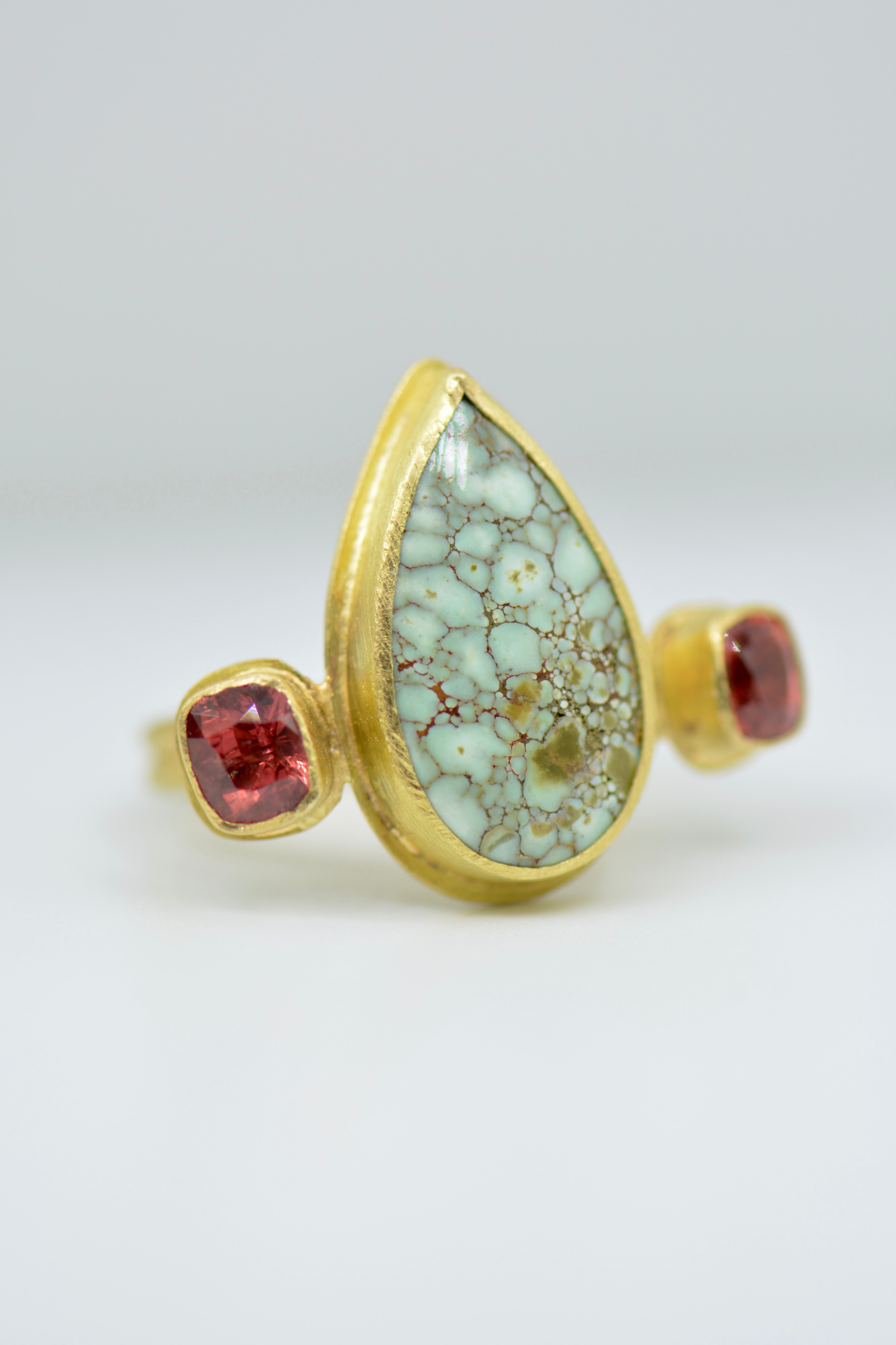 A gold ring featuring a large teardrop-shaped light green stone with a brown matrix pattern, flanked by two small faceted red gemstones, set against a neutral light gray background.