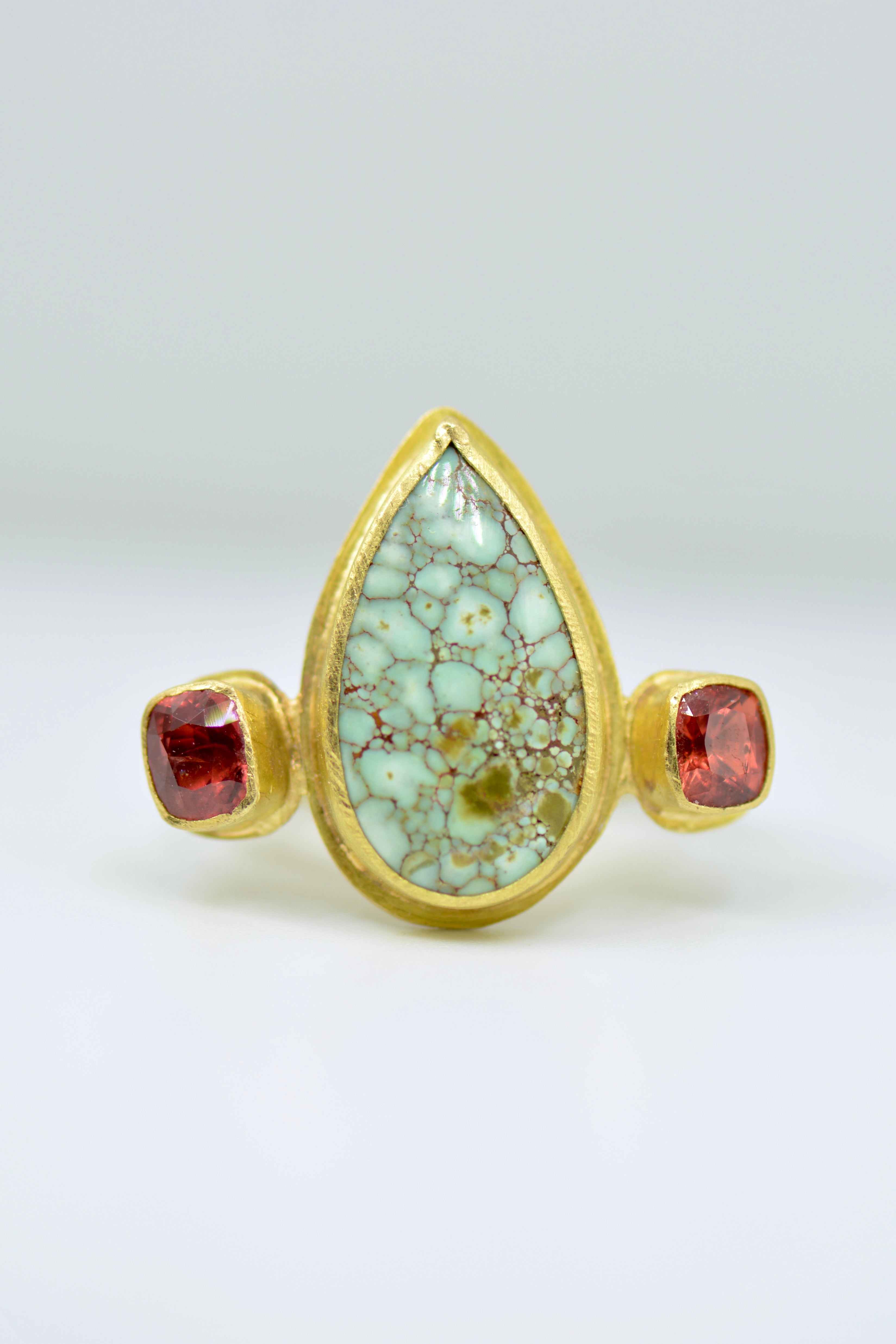 A gold ring featuring a large teardrop-shaped light green stone with an intricate brown matrix pattern, flanked by two small red cushion cut sapphire gemstones on a pale grey background.